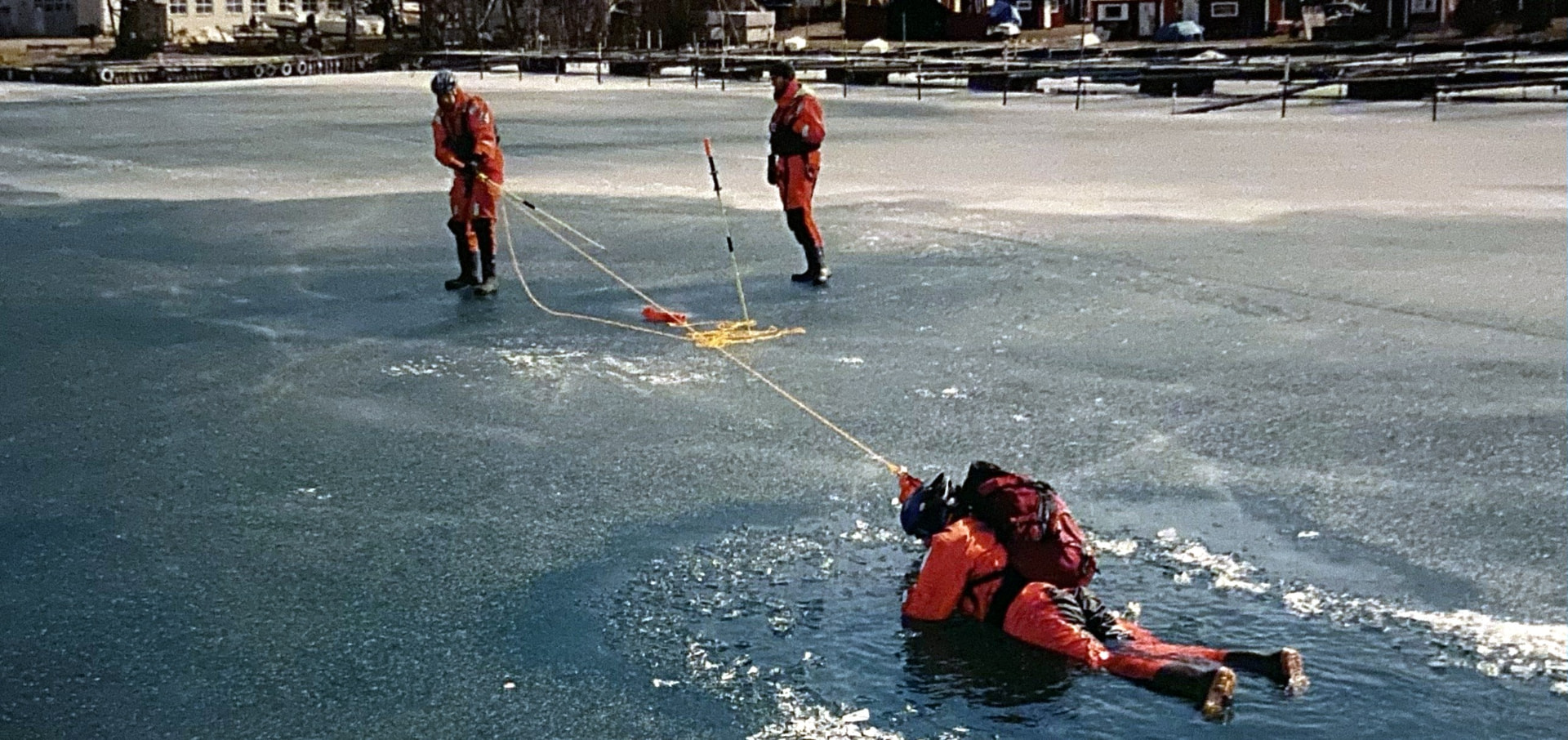 Ice rescue training: a person in a rescue suit in the water is pulled out by a line held by two skaters on the ice